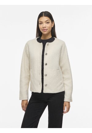 Nő krémszínű, gombos gyapjúkabátban fekete ing és fekete nadrág fölött, egyszerű fehér háttér előtt áll.