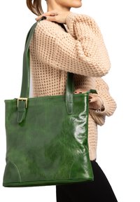 Groene leren totebag met een glanzende afwerking, rechthoekige vorm, dubbele handvatten en een gespaccent. Gedeeltelijk gedragen tegen een gestructureerde trui.