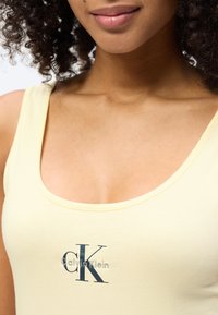 Γυναίκα με σγουρά μαλλιά που φοράει ανοιχτό κίτρινο τοπάκι με το λογότυπο Calvin Klein, χαμογελά απαλά με εμφανές γυαλιστερό κραγιόν στα χείλη.