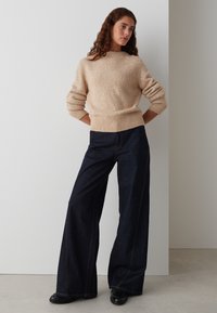 Pull en tricot beige avec motif texturé, encolure ronde et poignets côtelés ; associé à un pantalon en denim large bleu foncé et des bottines noires.