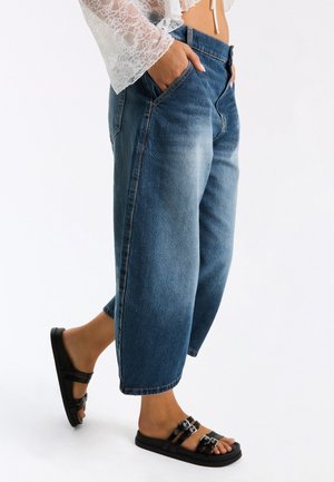 Vrouw draagt afgeknipte wijd uitlopende blauwe jeans, zwarte sandalen met gesp en een wit kanten top met handen in de zakken.