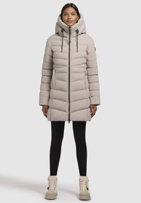 Manteau rembourré beige clair avec un col haut et une fermeture éclair. Dispose de sections matelassées horizontales, d'une capuche et de poches latérales. Assorti à des leggings noirs.