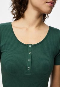 Camisola verde com textura canelada, apresentando um decote redondo e três botões de pressão prateados decorativos na parte da frente. Mangas curtas e textura suave.