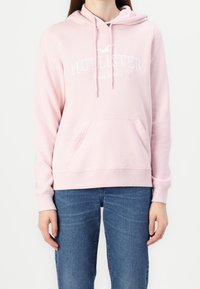 Osoba v světlém růžovém mikině Hollister s přední kapsou a modrými džíny, stojící na pozadí z čistě bílé barvy.