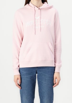 Personne portant un sweat à capuche rose clair de Hollister avec une poche avant et un jean bleu, se tenant contre un fond blanc uni.
