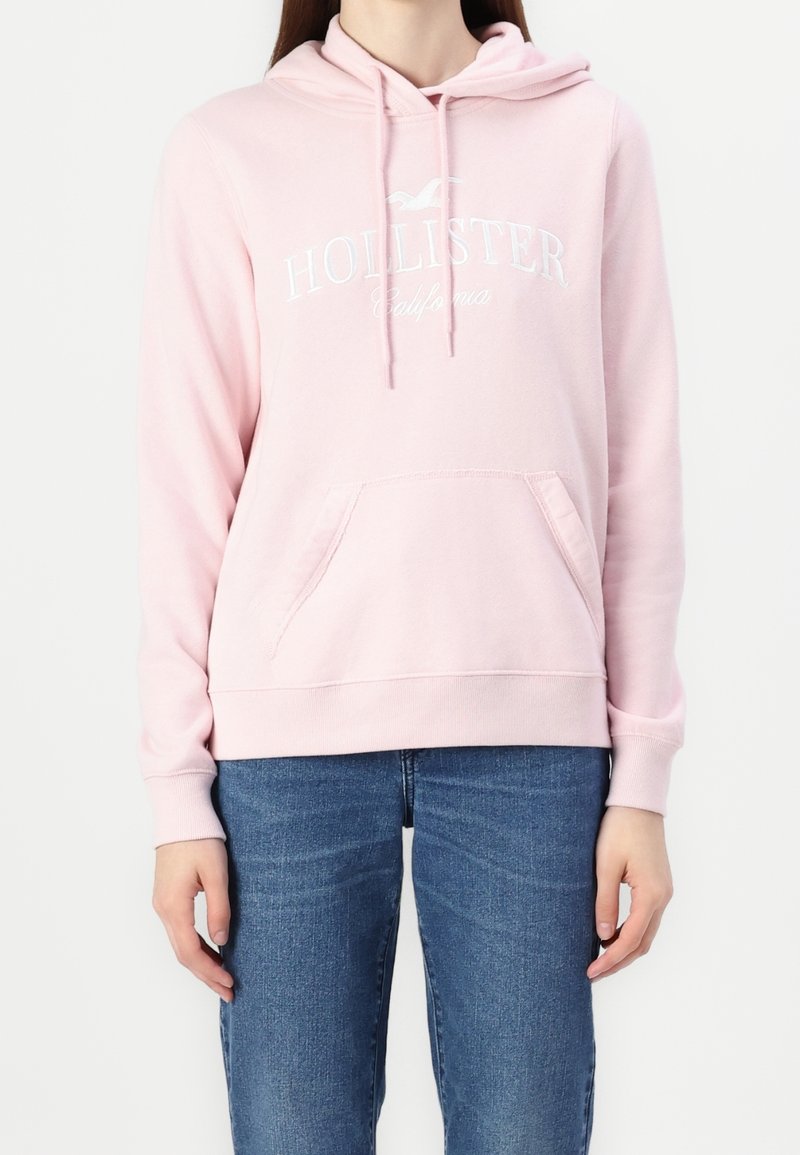 Osoba v světlém růžovém mikině Hollister s přední kapsou a modrými džíny, stojící na pozadí z čistě bílé barvy.