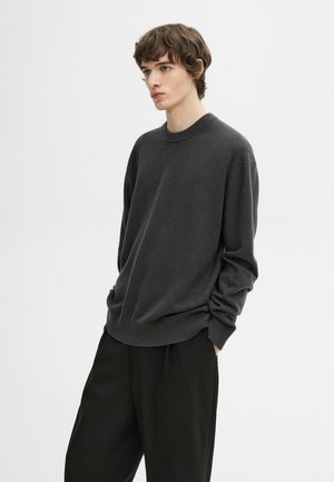Pull en tricot gris foncé avec un col rond, des manches longues et des poignets côtelés. Associe à un pantalon noir ample.