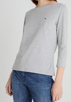 Femme portant un t-shirt manches longues gris clair avec un petit logo sur la poitrine et un jean bleu, montrée du cou jusqu'aux hanches.
