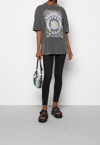 Camiseta gráfica gris de gran tamaño con estampado multicolor, leggings negros, sandalias gruesas negras y un pequeño bolso a rayas.