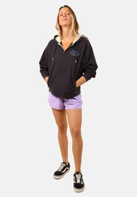 Haut à capuche noir zippé avec une capuche de couleur claire, arborant un logo multicolore ; associé à un short violet clair et des baskets noires.