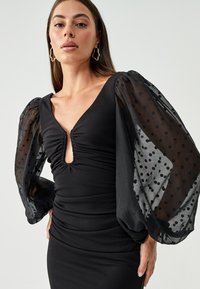 Black dress with a fitted bodice and puffy sheer sleeves featuring polka dots. V-neckline and gathered detailing at the center front.