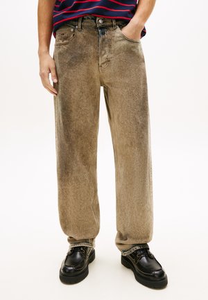 Mees, kes kannab lahtisi pruune pleekinud teksapükse, musti nahkkingi ja hoiab käed taskutes valge tausta ees.