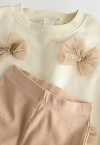 Creme sweatshirt med tre nudefarvede tylsløjfer, parret med ribbede bukser i blush pink. Stoffet ser blødt og hyggeligt ud, med et enkelt design.