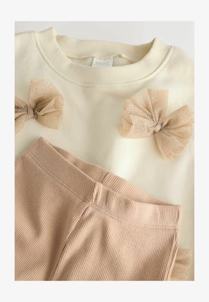Sweatshirt crème avec trois nœuds en tulle nude, associé à un pantalon rose pâle à côtes. Le tissu semble doux et confortable, avec un design simple.