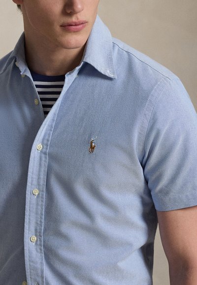 Vyras dėvi šviesiai mėlyną trumpomis rankovėmis marškinius su sagomis virš tamsiai mėlynos ir baltos dryžuotos apvalia apykakle marškinėlių su mažu siuvinėtu polo žaidėjo logotipu.