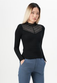 Must be black long-sleeve top with lace panel on the neckline, ribbed texture, and scalloped hem. Paired with blue trousers featuring side pockets.