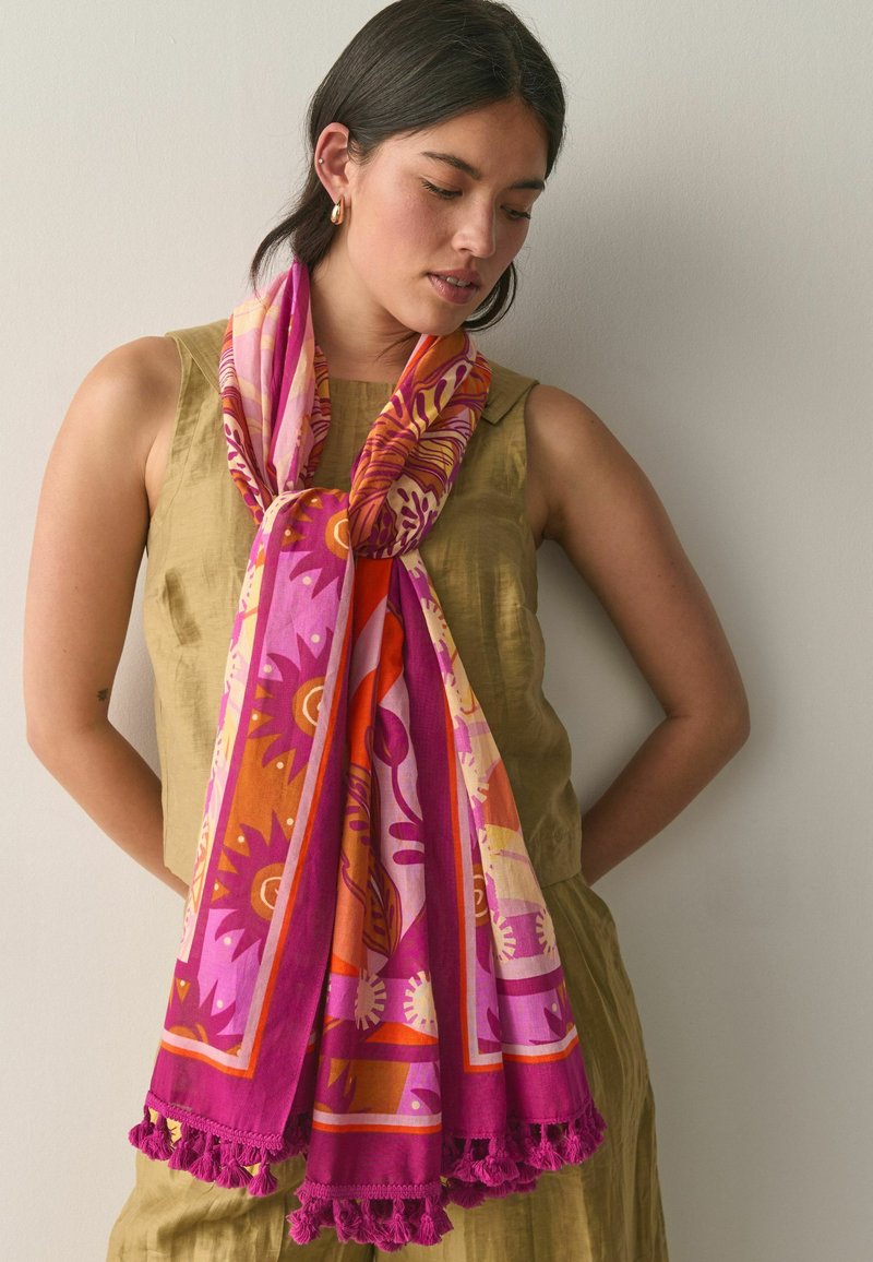 Femme portant une robe olive sans manches et une écharpe à motifs vibrants rose, orange et violet avec franges, debout contre un mur uni.