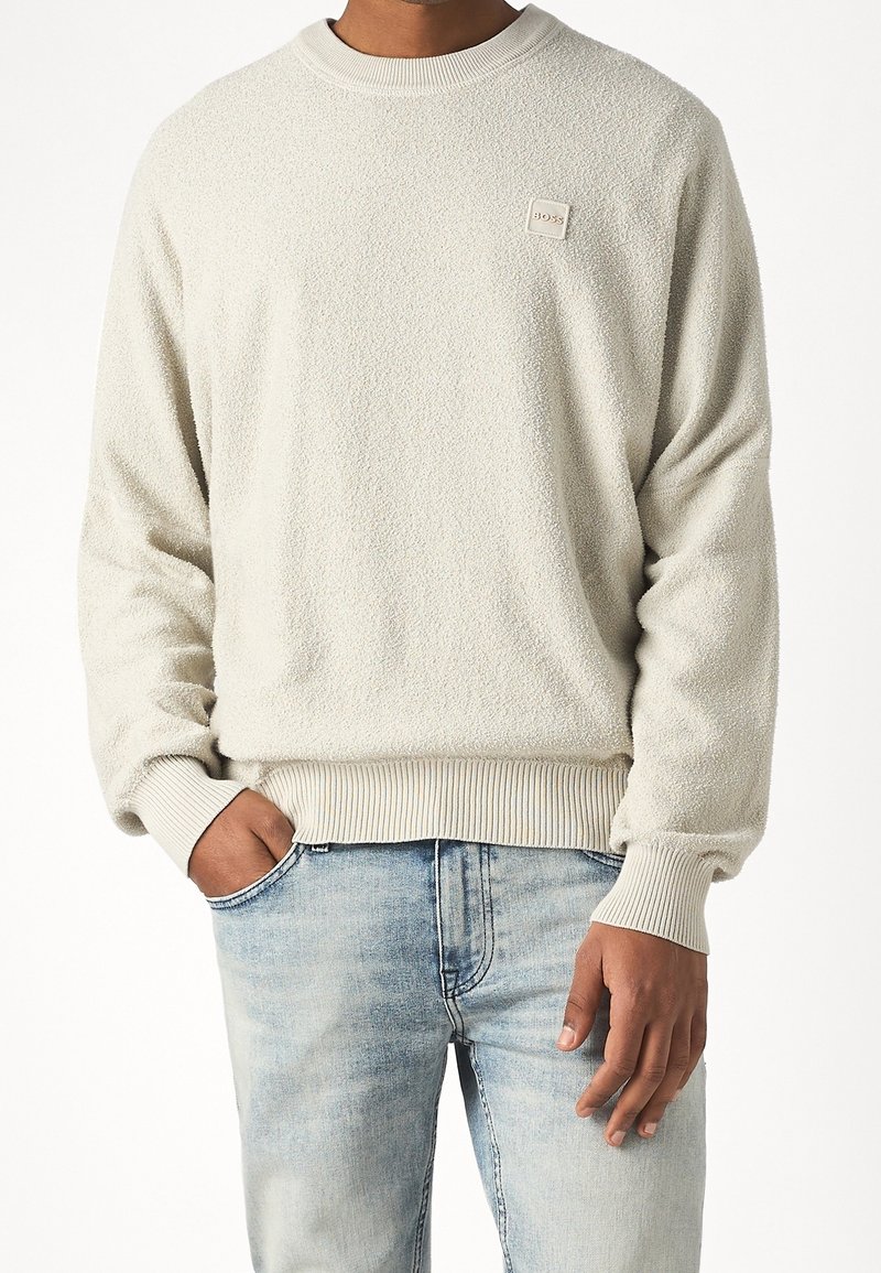 Homme portant un pull à manches longues texturé beige clair avec poignets et ourlet côtelés, associé à un jean bleu clair délavé, une main dans la poche.