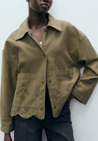 Veste vert olive avec col, dotée de poches avant et de détails brodés à l'ourlet. Fabriquée en tissu robuste avec une coupe décontractée.