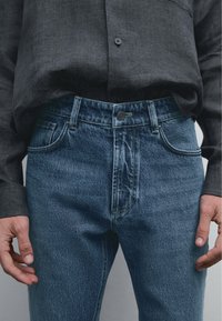 Denim jeans with a blue distressed finish, high waist, five-pocket design, and a black button closure, paired with a dark gray shirt.