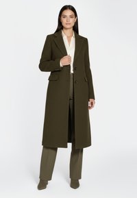 Lange, olijfgroene jas met een getailleerd ontwerp, een enkelrijige sluiting en twee zakken, gedragen over een crèmekleurige blouse en bijpassende broek.