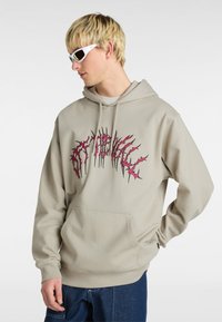 Lichtgrijze hoodie met een voorzak en een roze grafisch ontwerp dat lijkt op doornenachtige tekst. Model draagt witte zonnebril en marineblauwe jeans.