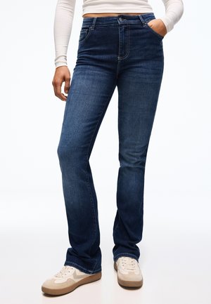 Personne portant un jean droit bleu foncé et des baskets beige, avec une main dans la poche et un haut blanc à manches longues raccourci à la taille.