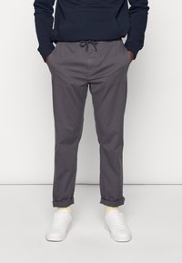 Grå bomullsbukser med elastisk midja och dragsnodd. Uppvikt byxben visar gula strumpor. Kombineras med vita sneakers.