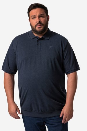 Mężczyzna z brodą w granatowej koszulce polo z krótkim rękawem i ciemnych dżinsach, stojący na białym tle.