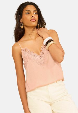 Tumejuustega naine kannab roosat pitsiga kaunistatud kamisooli, valgeid pükse, suuri rõngakujulisi kõrvaklambreid ja kolme jämedat käevõru randmel.