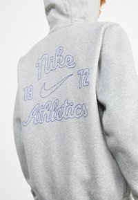 Felpa grigia con cappuccio, con testo ricamato blu: "Nike 19 12" e "Athletics." Texture morbida con polsini e orlo a coste.