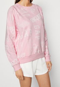 Modella che indossa un felpa rosa chiaro con i loghi "Barbie" e "GAP" in diverse dimensioni e direzioni, abbinata a pantaloni corti bianchi.