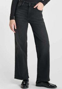 Zwarte wijde jeans met een hoge taille, gemaakt van textuurdenim. Voorzien van een knoopsluiting aan de voorkant en zakken, gecombineerd met glanzende zwarte instappers.