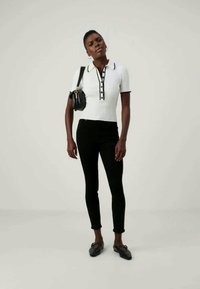 Álló nő, aki fehér, rövid ujjú, gombos pólót, fekete, testes nadrágot, fekete balerinacipőt visel, és egy fekete válltáskát hord.