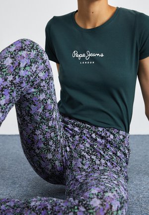 Osoba w ciemnozielonej koszulce Pepe Jeans London i fioletowych spodniach w kwiaty, siedząca z jedną nogą uniesioną na wykładzinie.