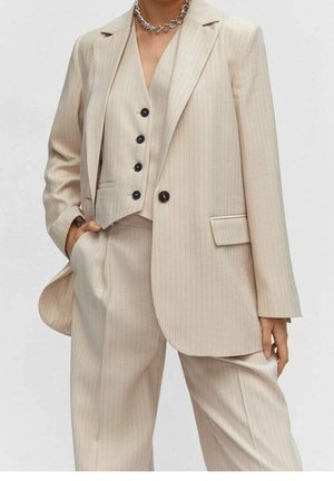 Vrouw draagt beige krijtstreep pak met blazer, bijpassend vestje met zwarte knopen, broek met wijde pijpen en zilveren ketting.