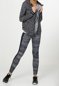 Woman wearing black and white patterned jacket, gray shirt, black and white patterned leggings, and black sneakers, standing with legs apart.