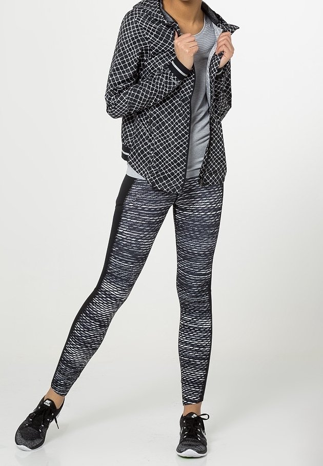 Woman wearing black and white patterned jacket, gray shirt, black and white patterned leggings, and black sneakers, standing with legs apart.