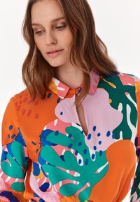 Färgglad blus i orange, rosa och grön med krage, som har abstrakta mönster och en nyckelhålsdetalj vid halsringningen, tillverkad av silkesliknande tyg.