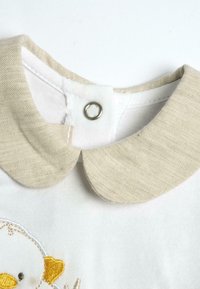 Camicia bianca con colletto Peter Pan in lino beige e chiusura a pressione. Presenta un ricamo di un'anatra gialla e un cuore.