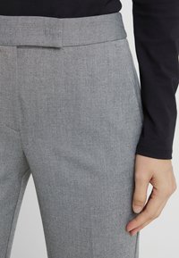 Close-up van een persoon in lichtgrijze gebreide broek en een zwart shirt met lange mouwen, met de rechterhand naast het lichaam.