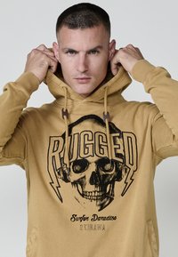 Beidzis hoody ar skeleta grafiku un vārdu "RUGGED" treknos burtos. Iekļuvis ar aukliņu kapuci un kanguru kabatu.