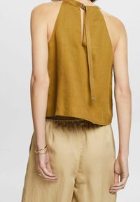 Top sin mangas de color mostaza con espalda con abertura en forma de llave, lazo ajustable y dobladillo curvado. Combinado con pantalones holgados beige.