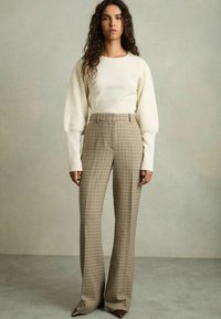 Off-white stickad tröja med långa, något puffiga ärmar, parat med högmidjade, rutiga bruna och beige byxor samt spetsiga bruna skor.