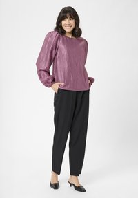 Texturierte, pflaumenfarbene Bluse mit Puffärmeln, kombiniert mit lockeren schwarzen Hosen. Das Model steht mit den Händen in den Taschen und trägt schwarze Mules.