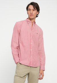 Young man wearing a red and white striped button-up shirt and beige pants, standing with one hand in his pocket, looking to the side.