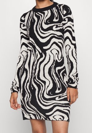 Kobieta w dzianinowej sukience z długim rękawem w czarno-biały abstrakcyjny wzór wiru, z okrągłym dekoltem.