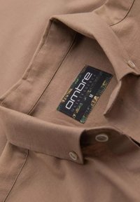 Chemise beige à col en tissu doux. Porte une étiquette avec la marque "ombre", deux boutons et une texture lisse.