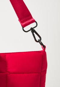 Sac matelassé rouge présentant une surface texturée, une sangle réglable et un clip en métal noir pour le sécuriser.