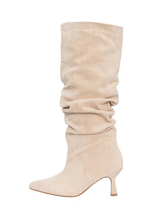 Bota holgada de ante beige hasta la mitad de la pantorrilla, con punta puntiaguda y tacón kitten corto, vista de perfil sobre un fondo blanco.